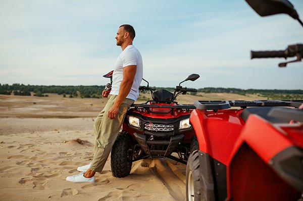Brutal man poses near atv in desert, quadbike Brutal man poses near atv in desert sands. Male person on quad bike, sandy race on quadbike, dune safari in hot sunny day, 4x4 extreme adventure, quad-biking